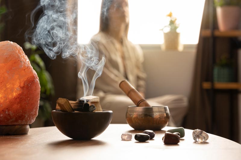 Palo Santo, Klangschale und Steine stehen auf einem Tisch. Es ist eine wohlig warme Atmosphäre, im Hintergrund sieht man eine Person sitzen.